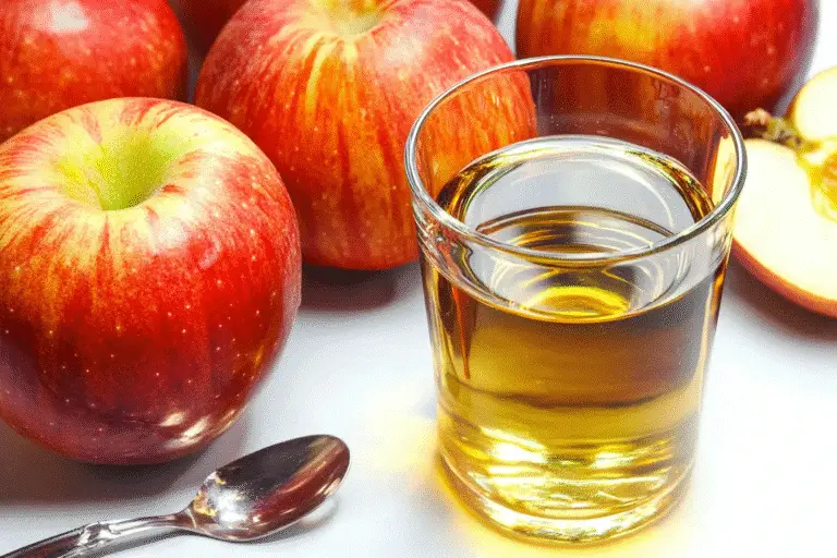 A glass of apple cider vinegar surrounded by fresh red apples and a metal spoon on a white surface.