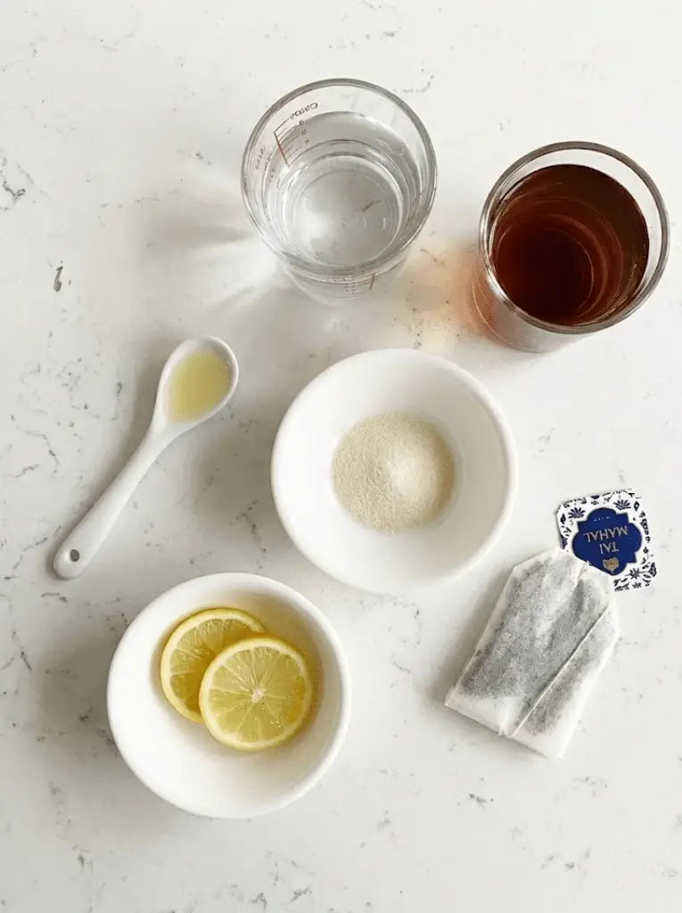 Gelatin trick recipe ingredients - unflavored gelatin powder, tea, water, lemon slices, and natural sweetener arranged on white marble counter